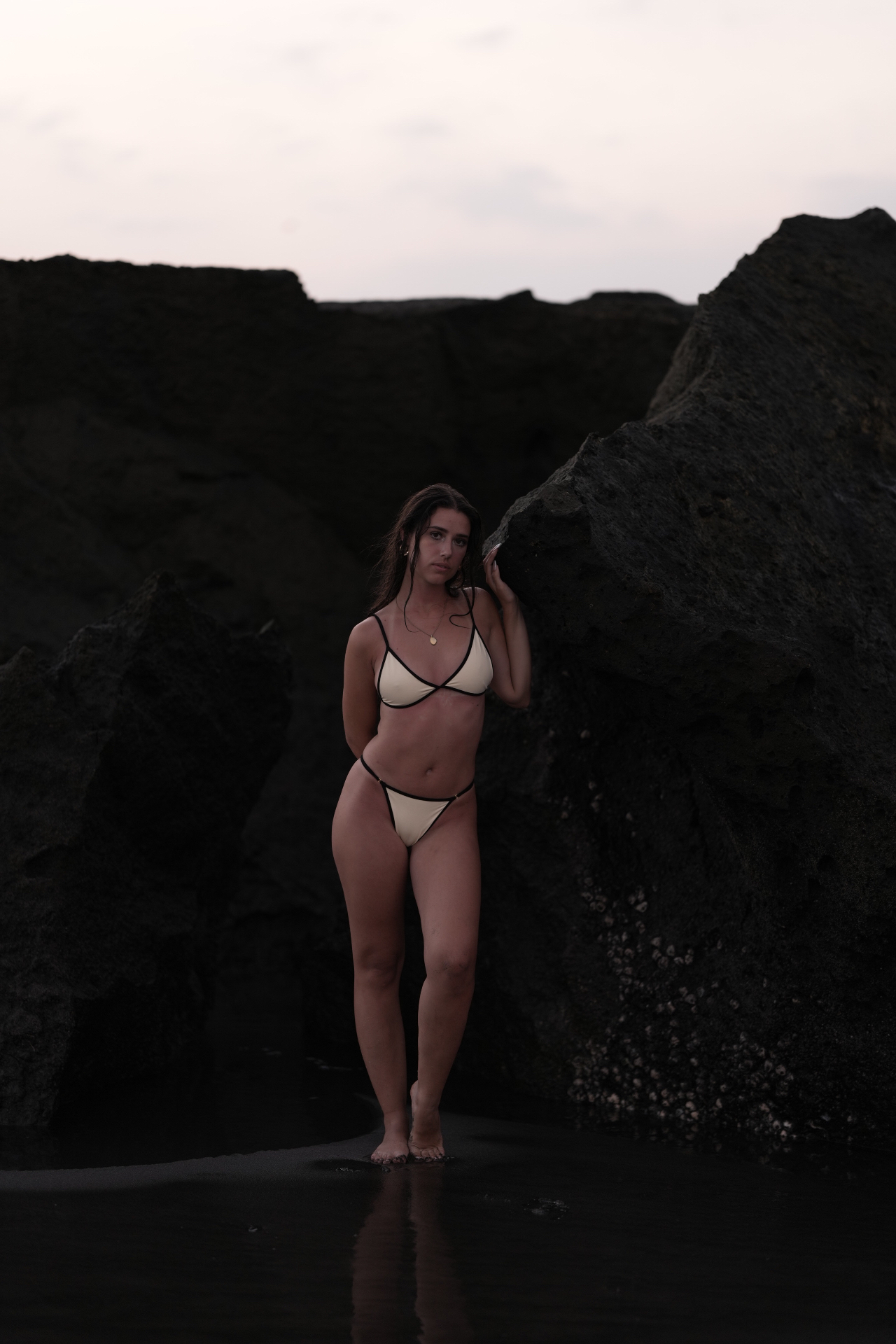 Model standing between volcanic rocks at dusk wearing the PAGI Essential bikini in ivory with black trim